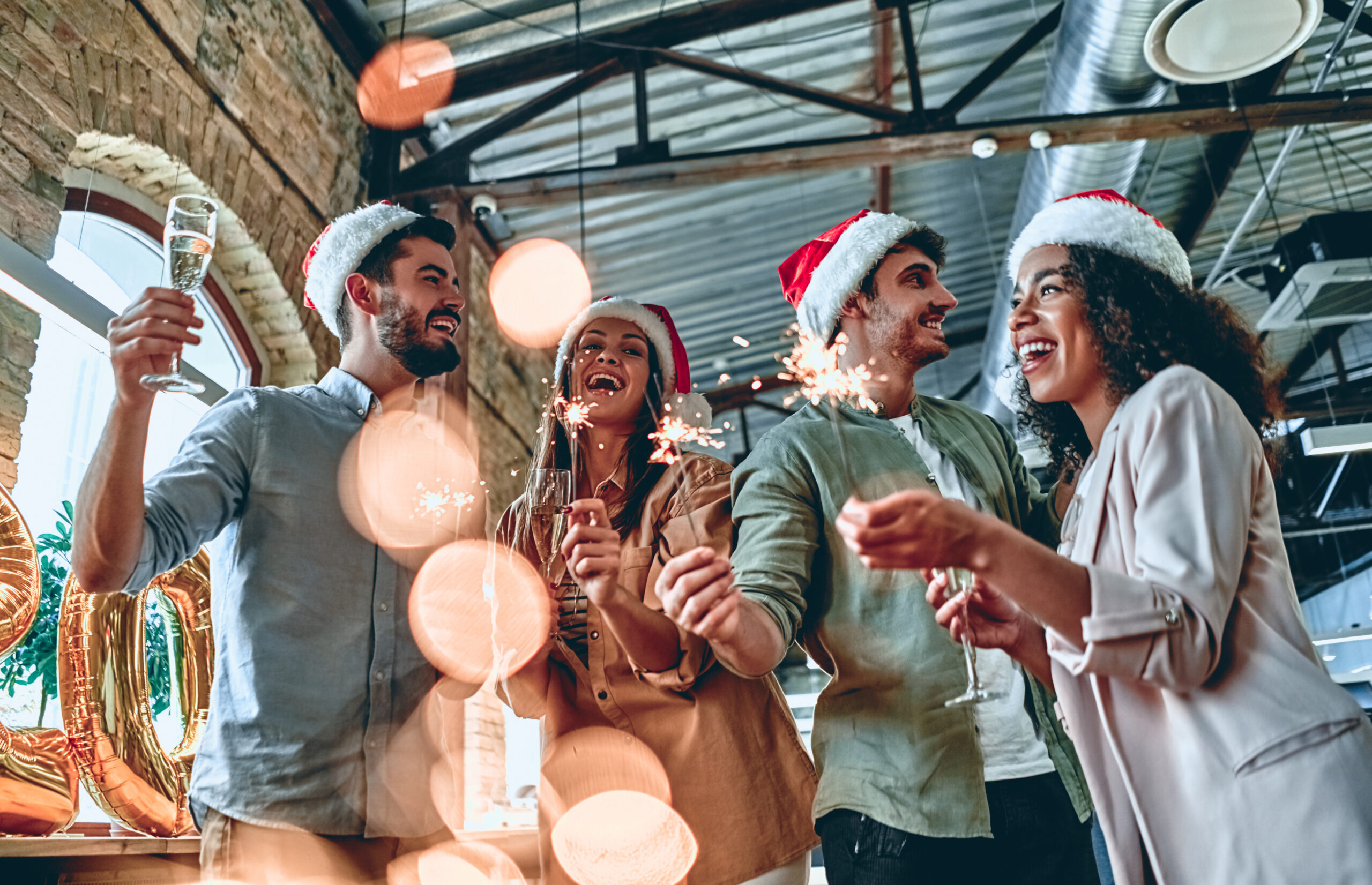 Multiracial young creative people are celebrating holiday in modern office. Group of young business people are drinking champagne with sparkling bengal lights in coworking. Successful hipster team of freelancers.