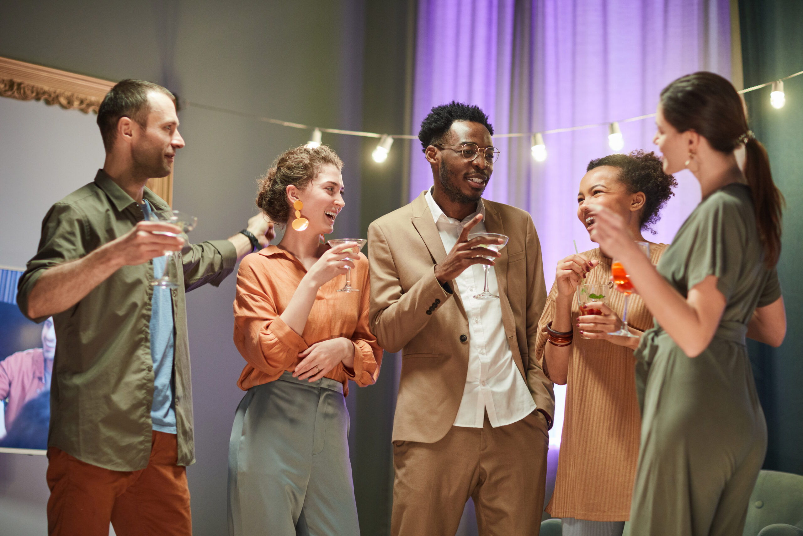 Waist up portrait of diverse group of friends having fun and drinking cocktails at home party